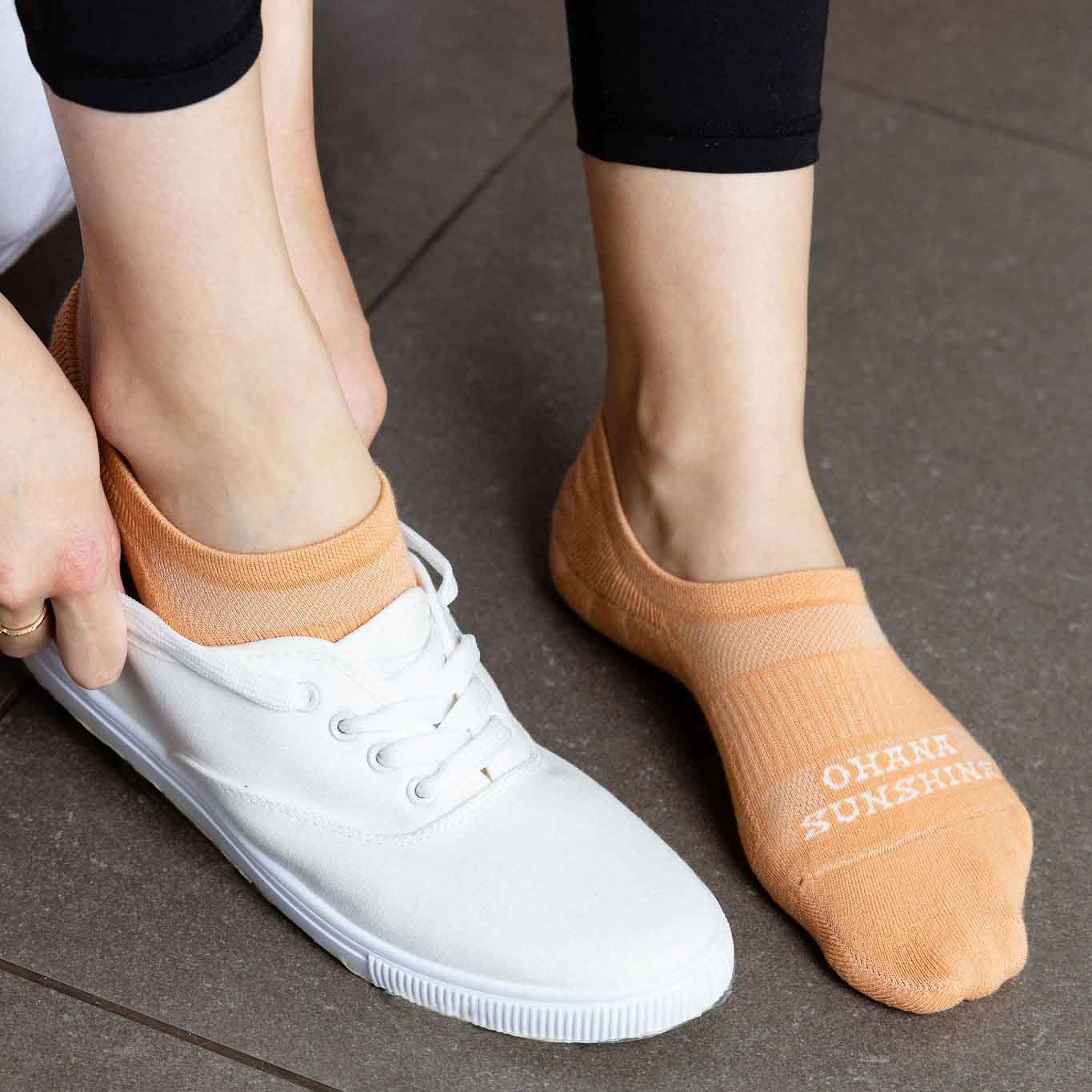 Female model wearing orange Ohana Sunshine no-show socks, with one foot slipping into a white sneaker to show how the socks stay hidden and secure
